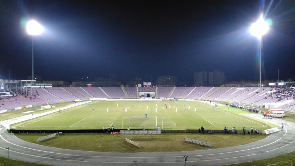 Two tall floodlights shine over the pitch in an almost empty football stadium