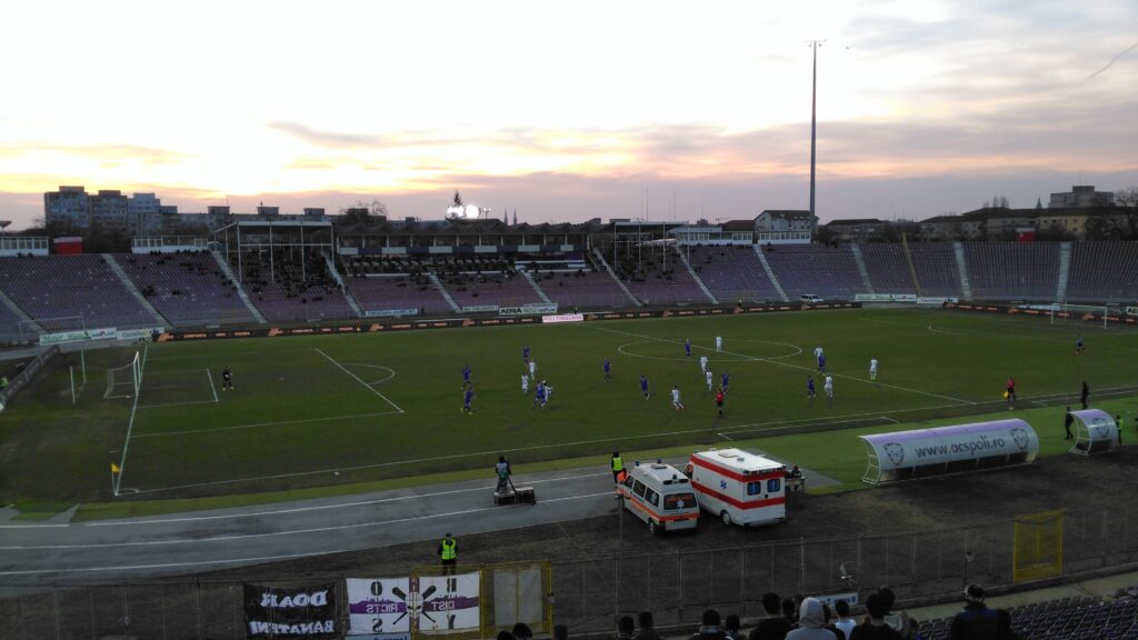 A football match in progress in a large football stadium, two ambulances are parked near the perimeter of the pitch